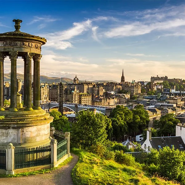 A scenic view of the Cliffs of Moher in Ireland, showing dramatic, steep cliffs rising above the Atlantic Ocean with lush green grass on top and sunlight illuminating the rugged rock faces. The sea below is calm with gentle waves.