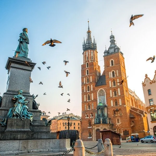 A historic European square with a large brick cathedral featuring twin towers in the background, a statue of a man with sculpted figures at the base, and birds flying in a clear sky.