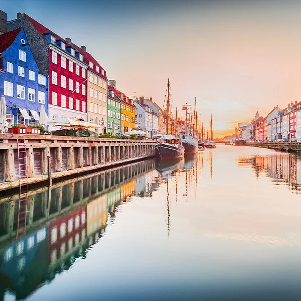 Colorful historic buildings line the waterfront of Nyhavn in Copenhagen, Denmark, with boats moored along the canal and their reflections visible in the calm water at sunset.
