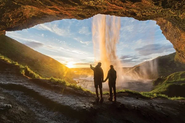 Two people stand hand in hand behinda a large waterfall, framed by a rocky overhang, with sunlight streaming through the falling water and a dramatic sky in the background.