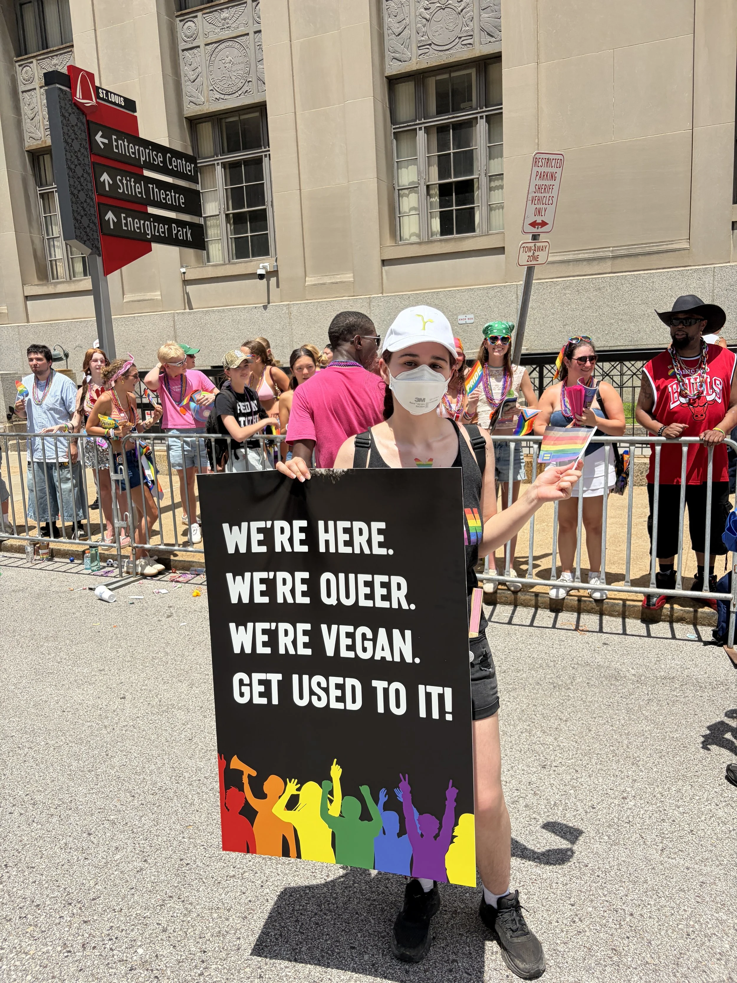 A queer vegan activist holding a signs that says we_re here, queer, and vegan in front of a crowd at St. Louis PrideFest.jpg