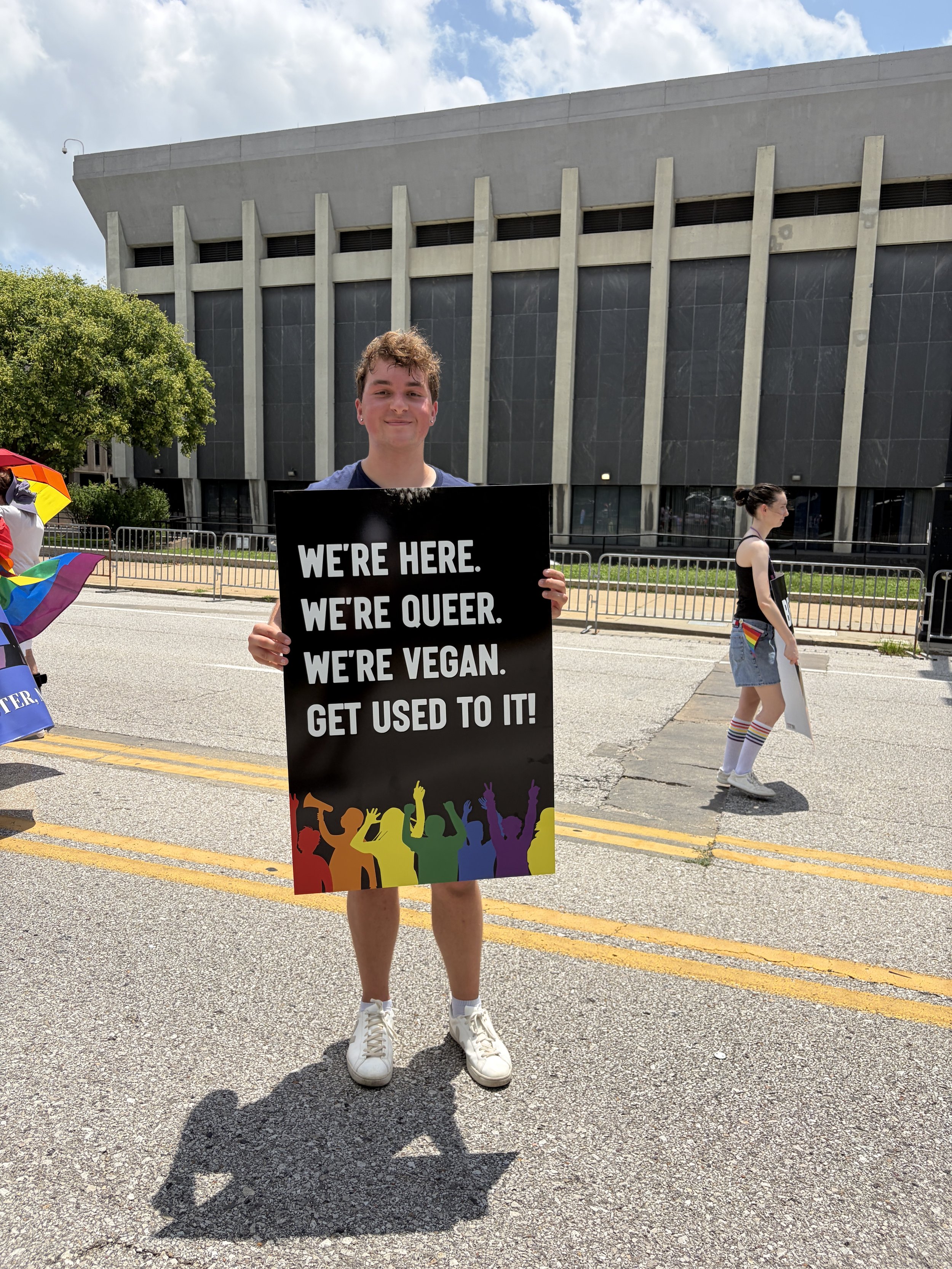 Troy proudly posing with his sign that reads we_re here, queer, and vegan at our Vegan Pride Parade.jpg