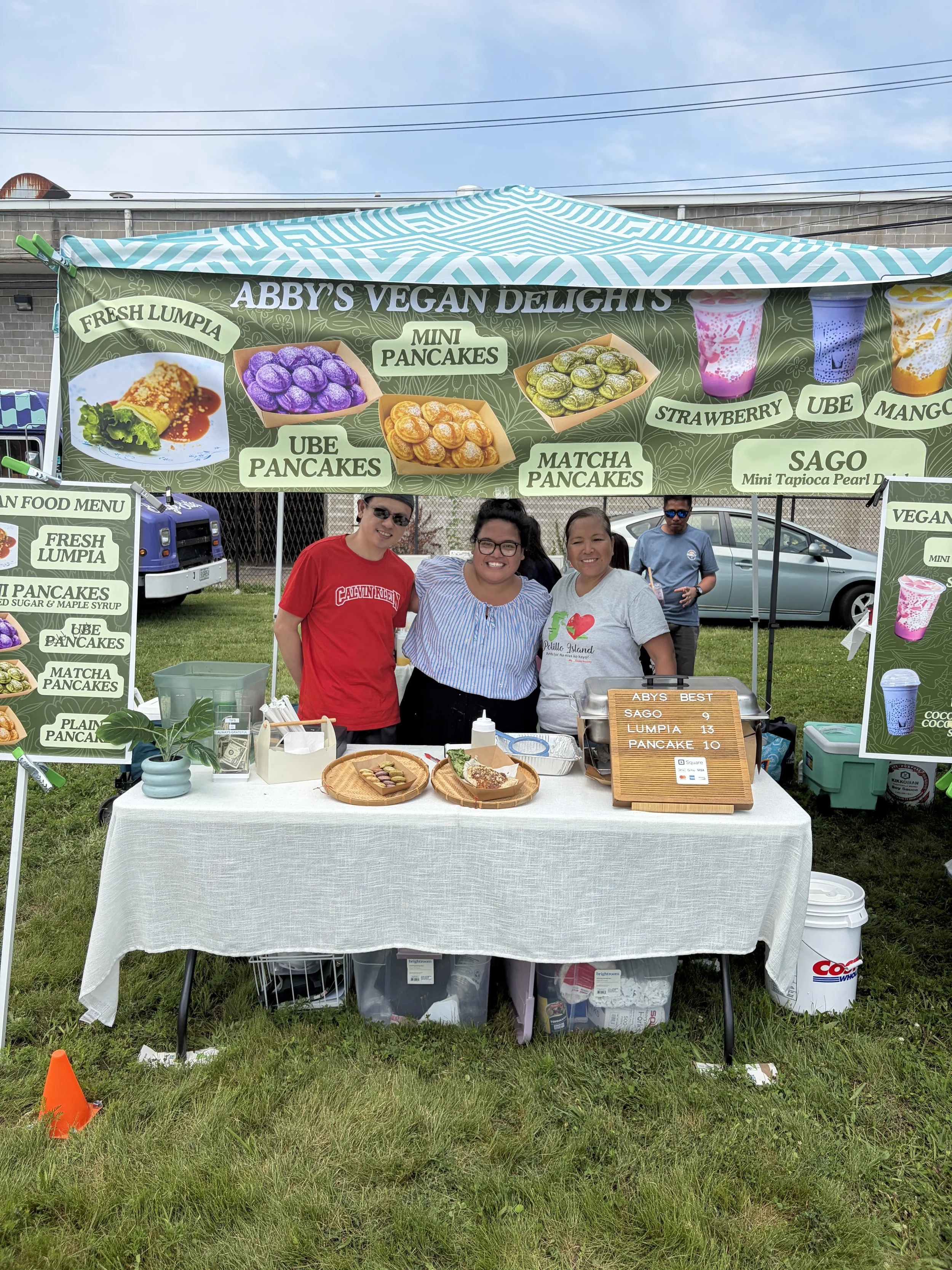 Owner and employees of Abby_s Vegan Delights pose behind booth at STL Vegan Market in St. Louis, Missouri.png