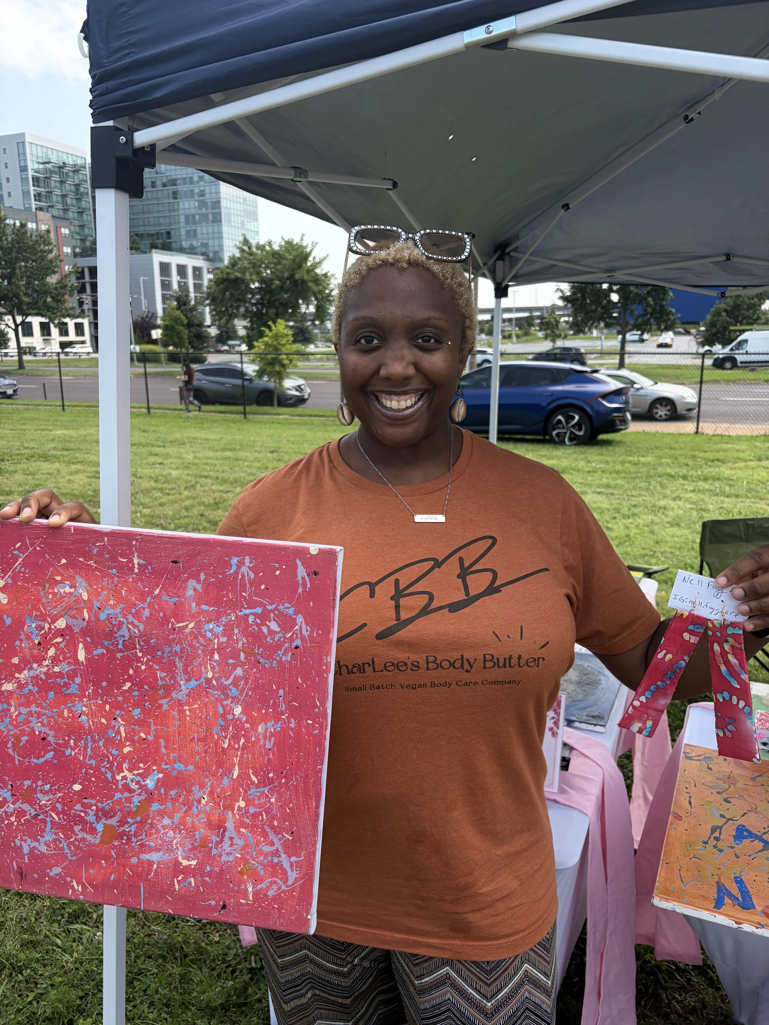Vendor of Charlee_s Body Butter poses with colorful artwork at STL Vegan Market in St. Louis, Missouri.png