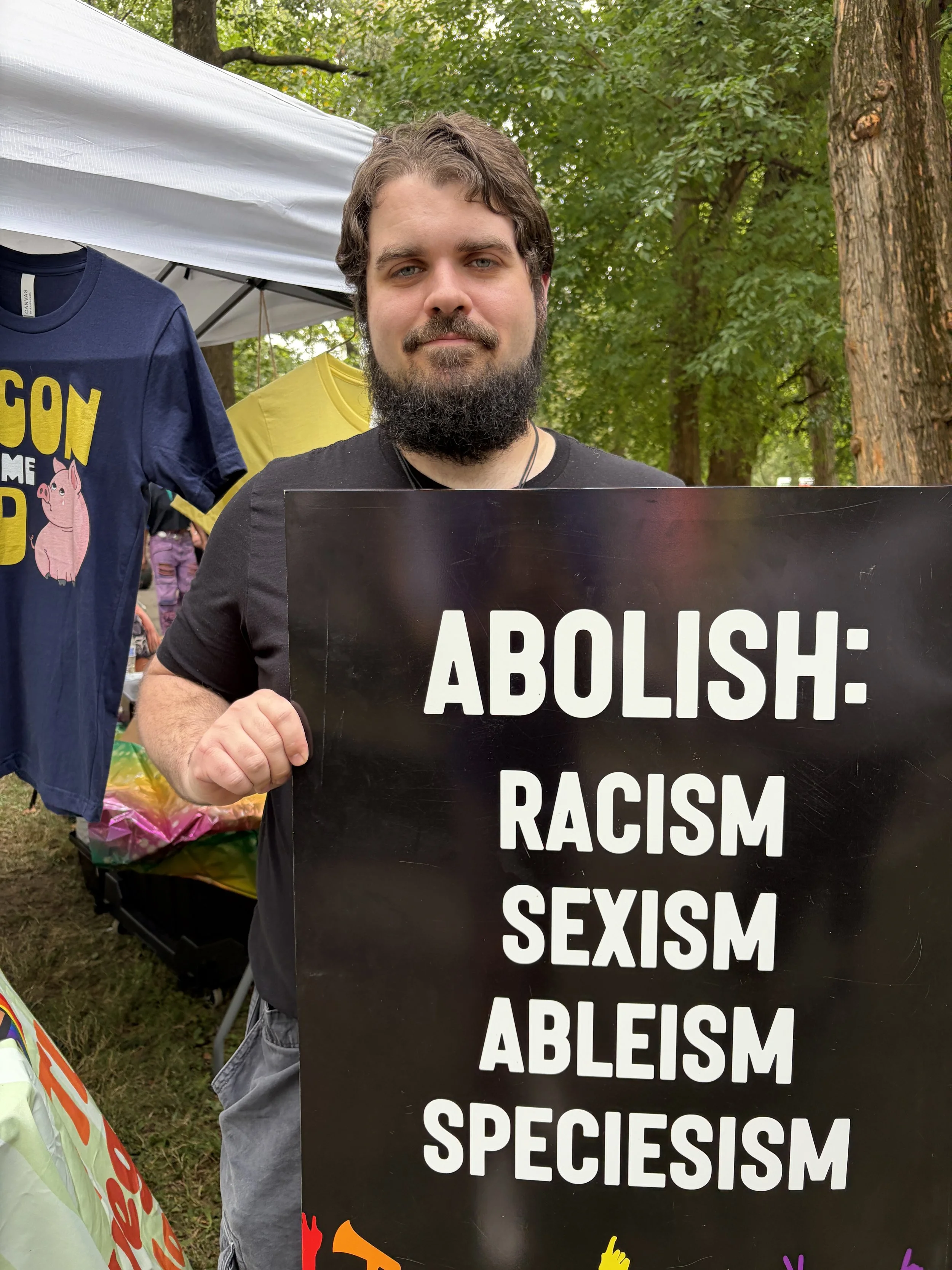Alex happily holds sign for Vegan Pride Booth at Tower Grove Pride in St. Louis, Missouri.png