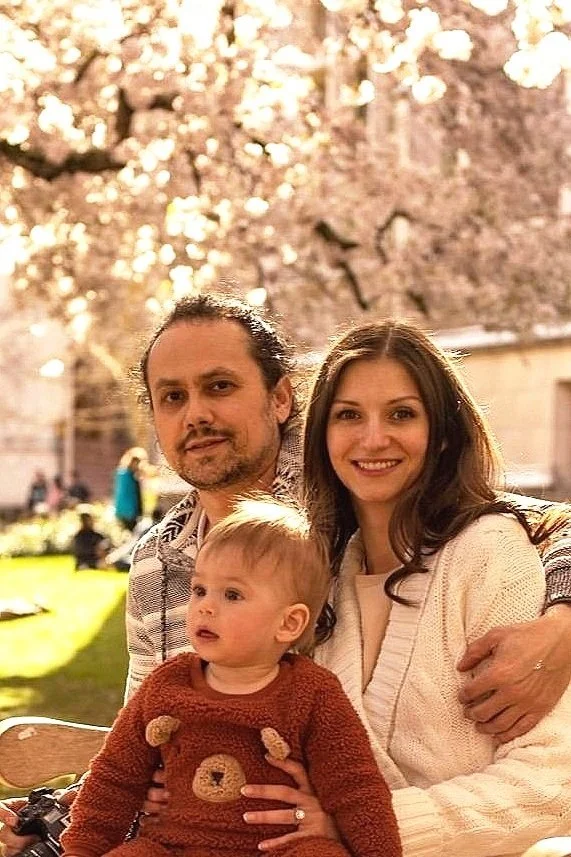 A family portrait outdoors with cherry blossoms; a man, woman, and child sitting closely.