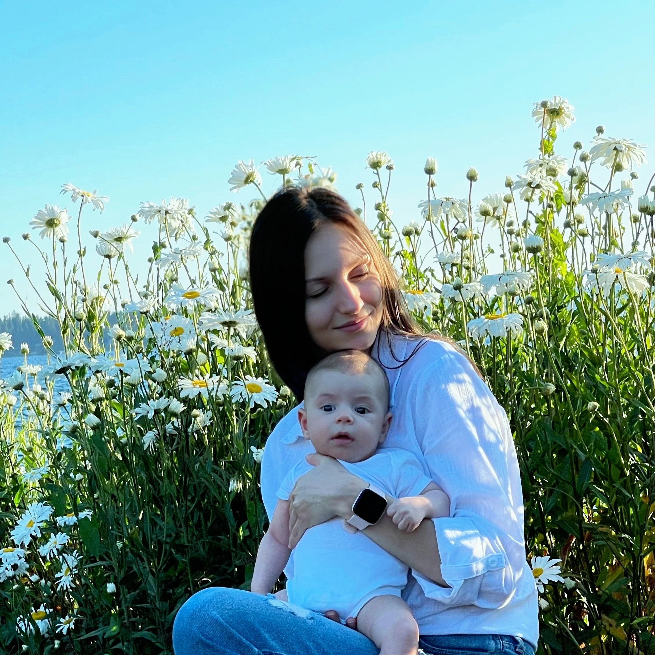 A woman holding a baby sits amidst a field of daisies with a clear blue sky in the background.