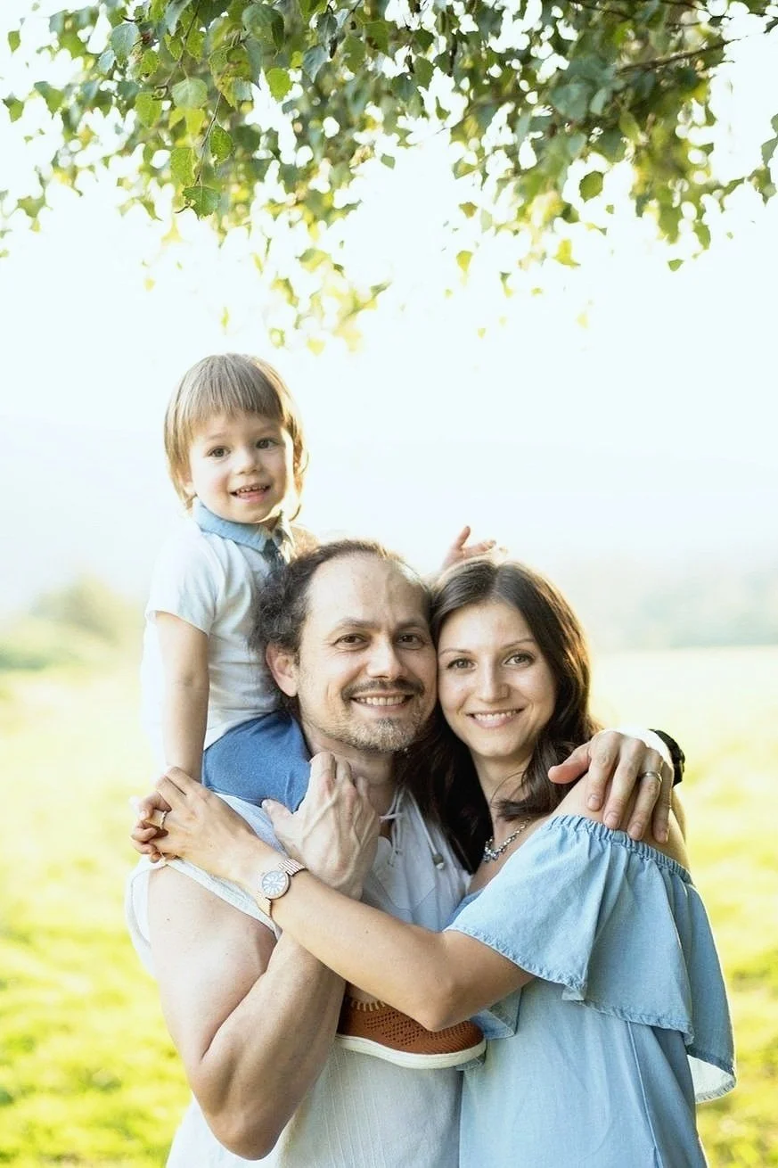 A family of three outdoors, smiling and hugging. A man and woman are holding a child on the man's shoulders. Trees in the background.