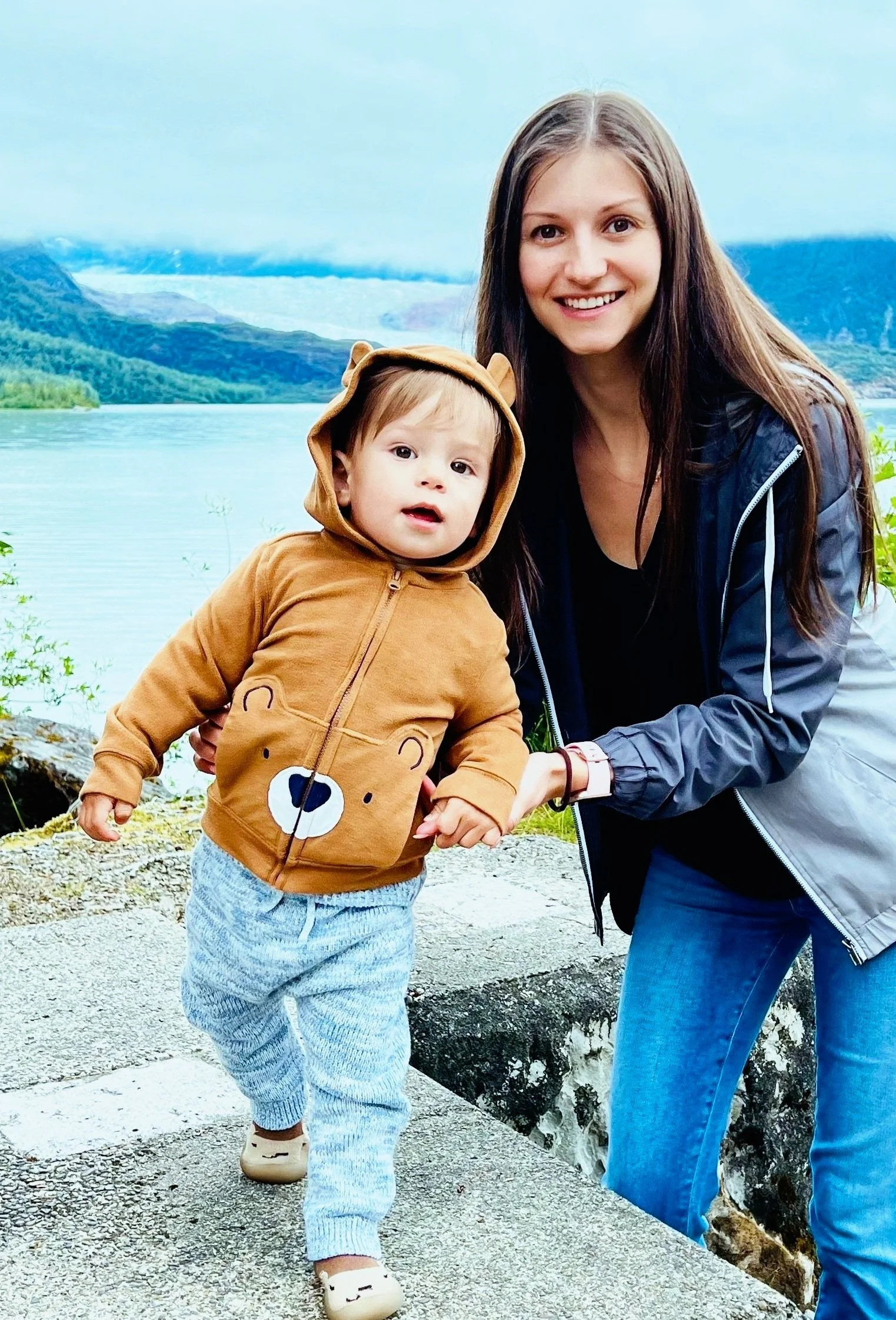 A woman and a child outdoors near a scenic lake with mountains in the background. The child is wearing a brown hoodie with bear ears and design, blue pants, and beige shoes.