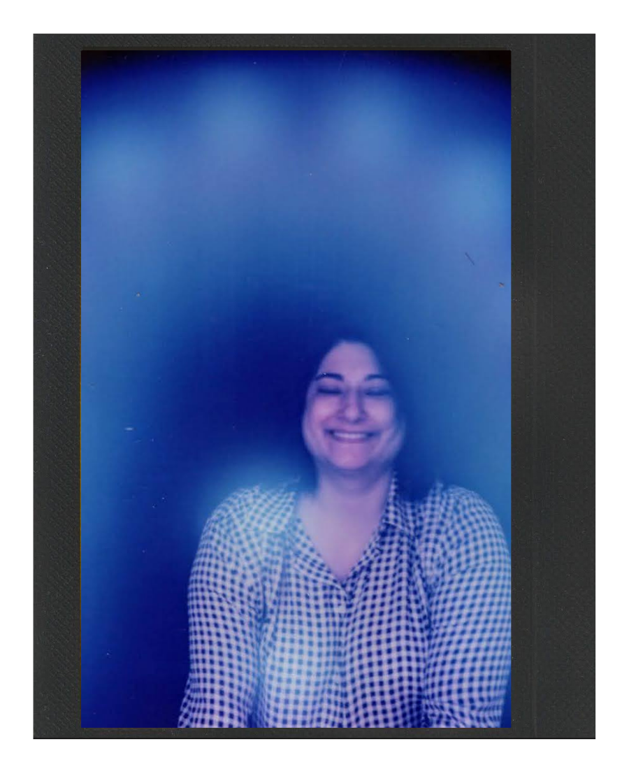 A woman with dark hair and a checkered shirt smiling with eyes closed, captured in a dimly lit environment with a blue haze.