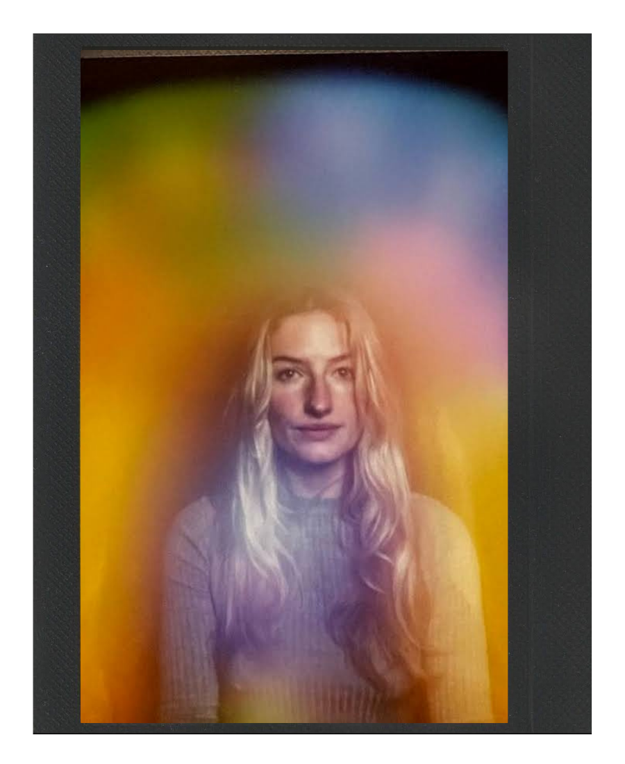 A woman with long blonde hair and a beige ribbed sweater standing against a colorful blurred rainbow background.