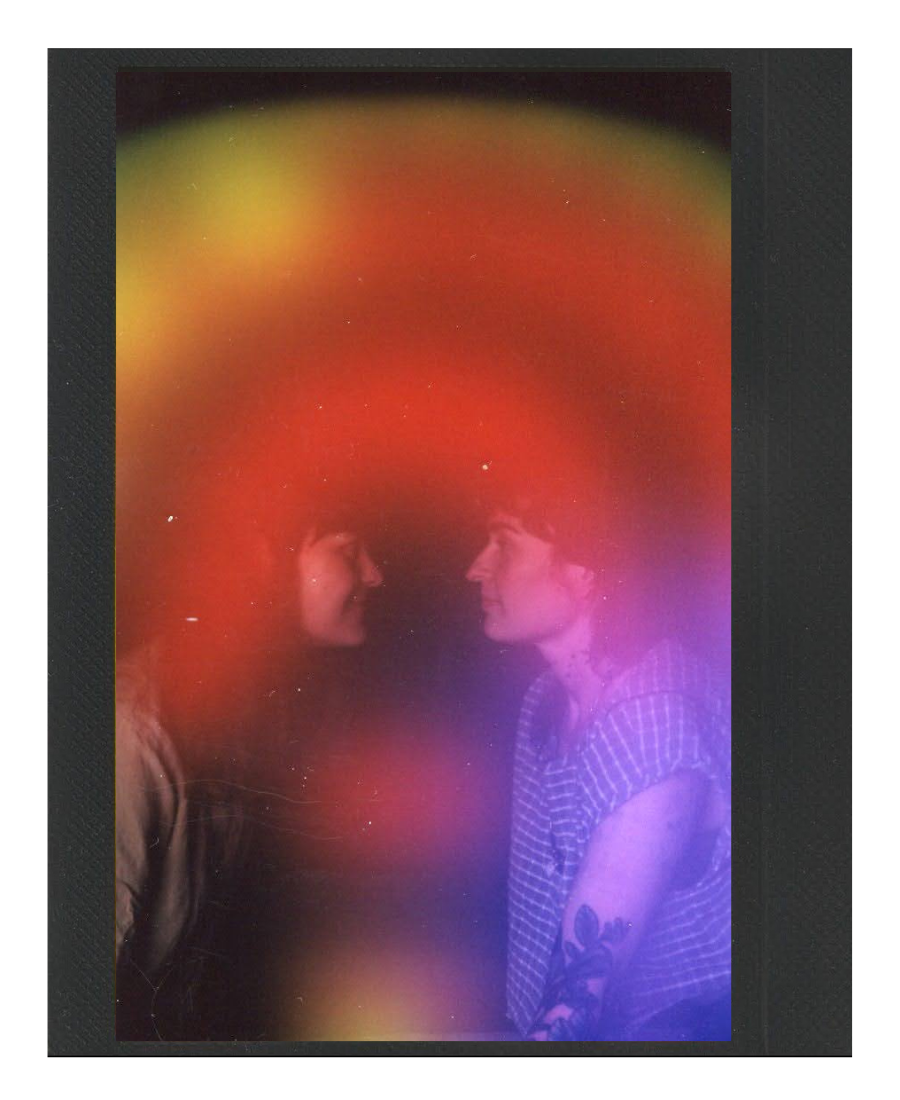 Two women face each other in a dark setting with colorful, blurry lights surrounding them, creating a dreamy, intimate atmosphere.