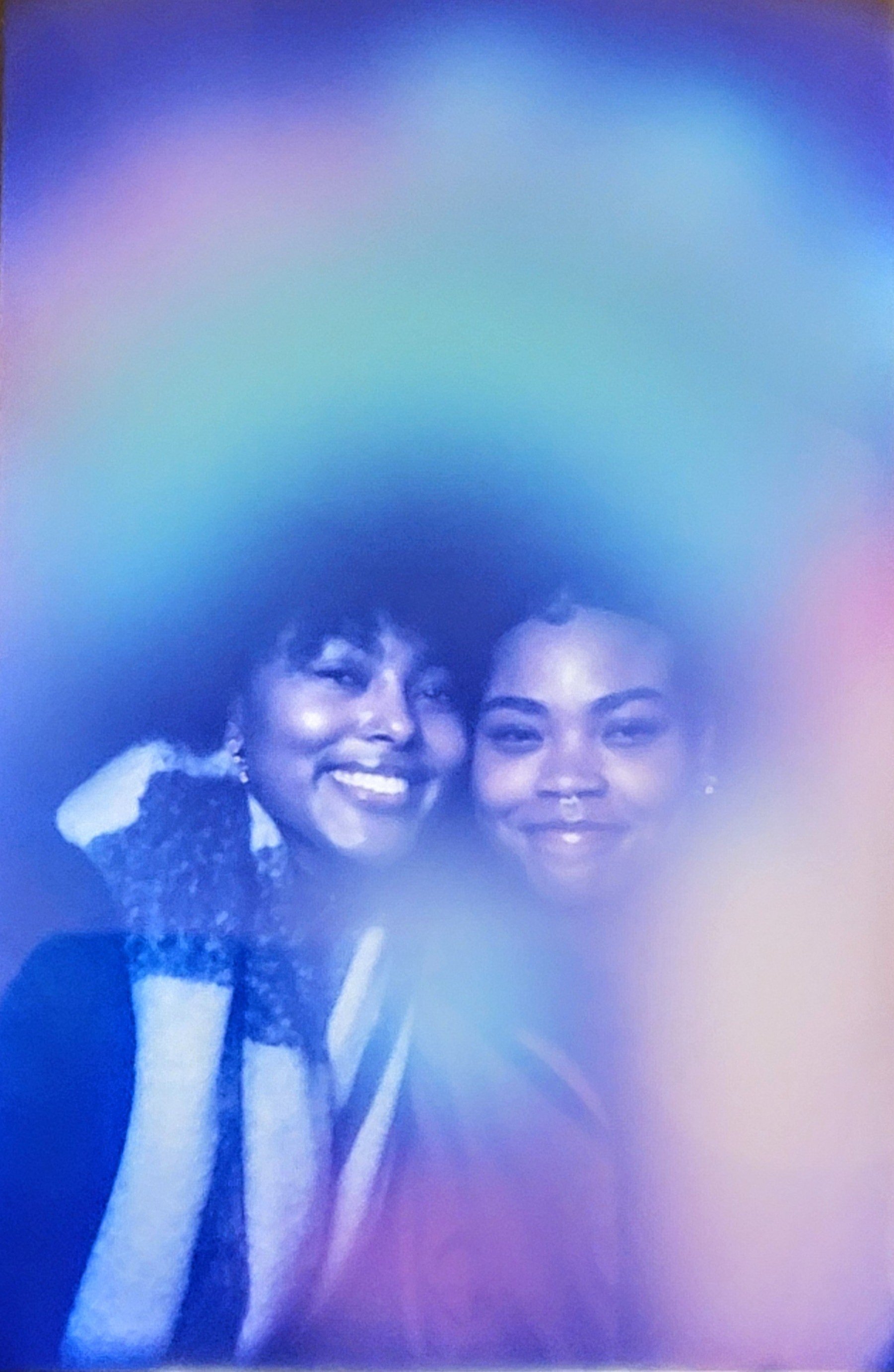 Two women smiling, one with short dark hair and the other with long dark hair, close together in a photo with a colorful, blurred background.
