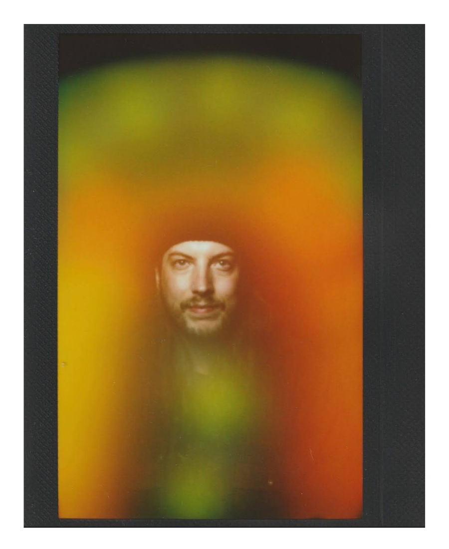 A man's face is visible through a colorful, blurry surface with patches of green, yellow, orange, and red, resembling a fruit.
