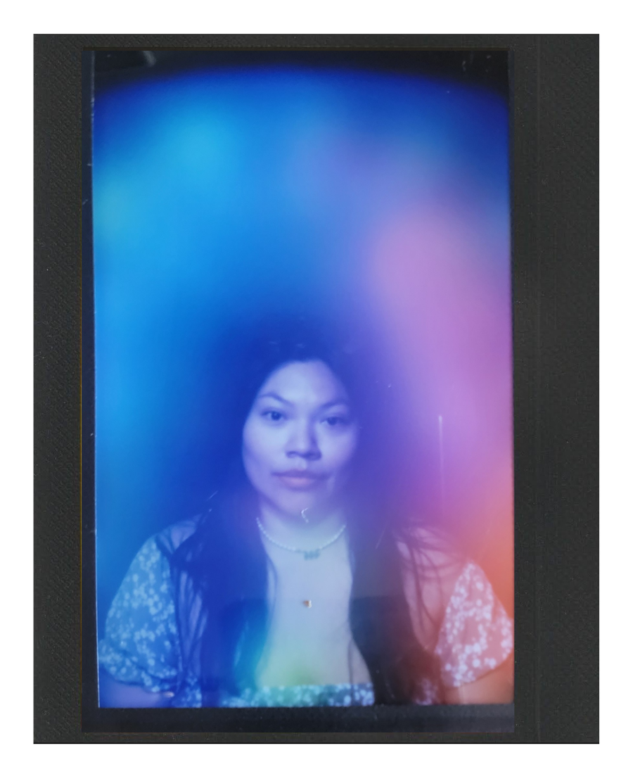 A woman with long dark hair and wearing a floral top, necklaces, and a choker, looking at the camera with a neutral expression, displayed on a screen with colorful light effects.