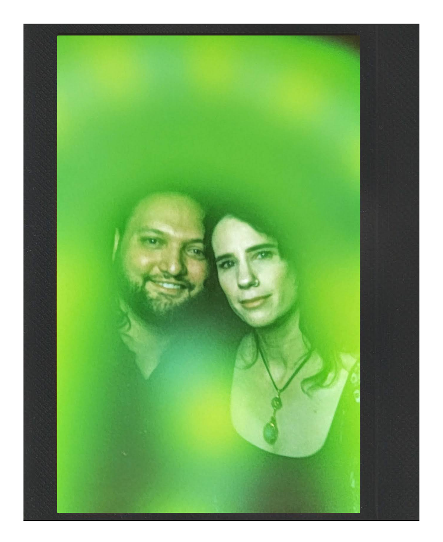 A man and woman taking a selfie together through a green tinted glass or screen, both smiling with the woman wearing a necklace and a nose piercing.
