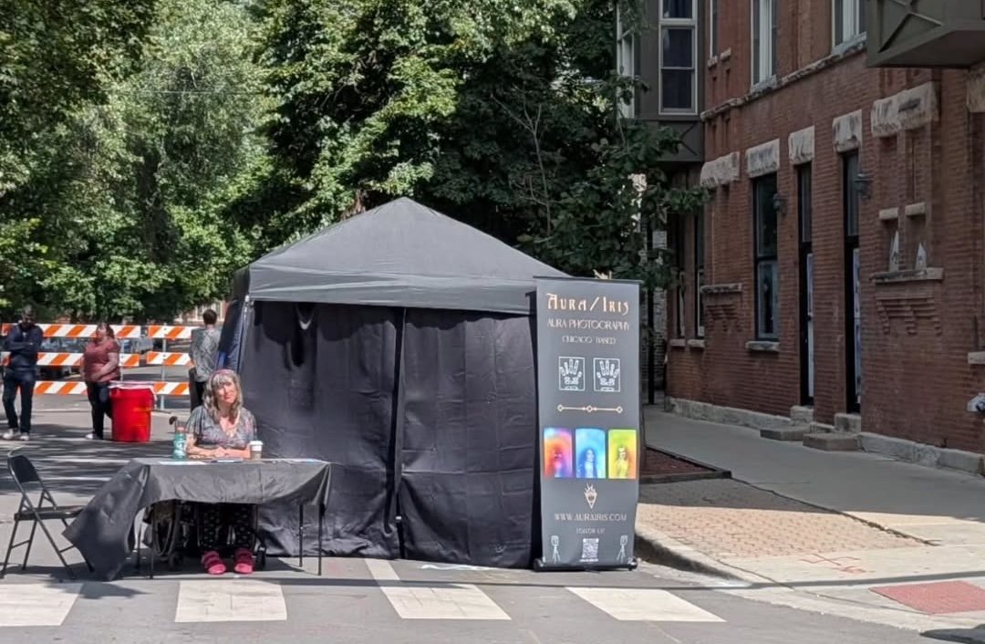 Outdoor booth with a grey canopy and a black table, woman sitting at the table, sign with colorful images and website URL, brick building and trees in background.