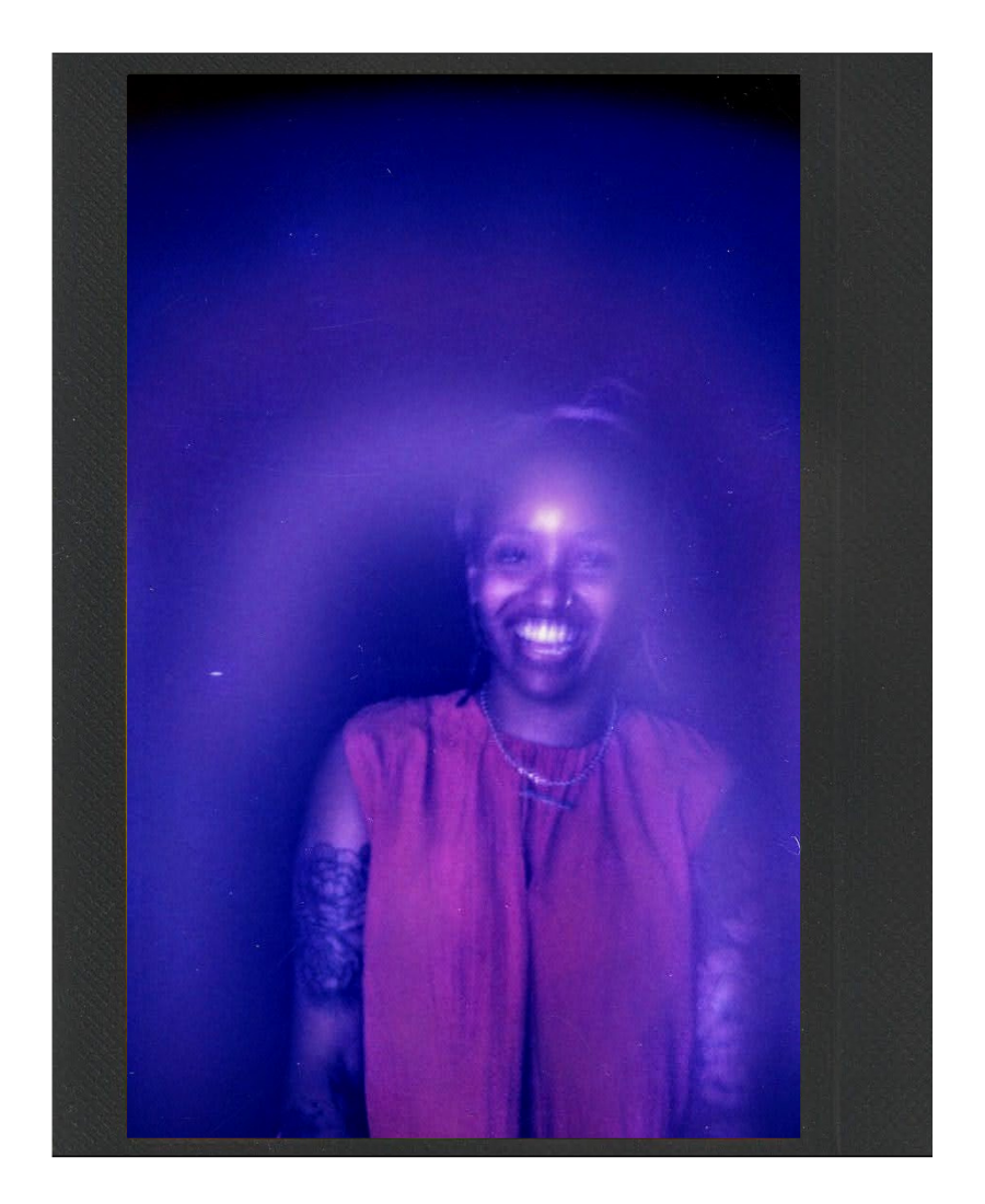 A woman with a bright smile, wearing a sleeveless red top and necklaces, standing against a dark background with purple lighting.