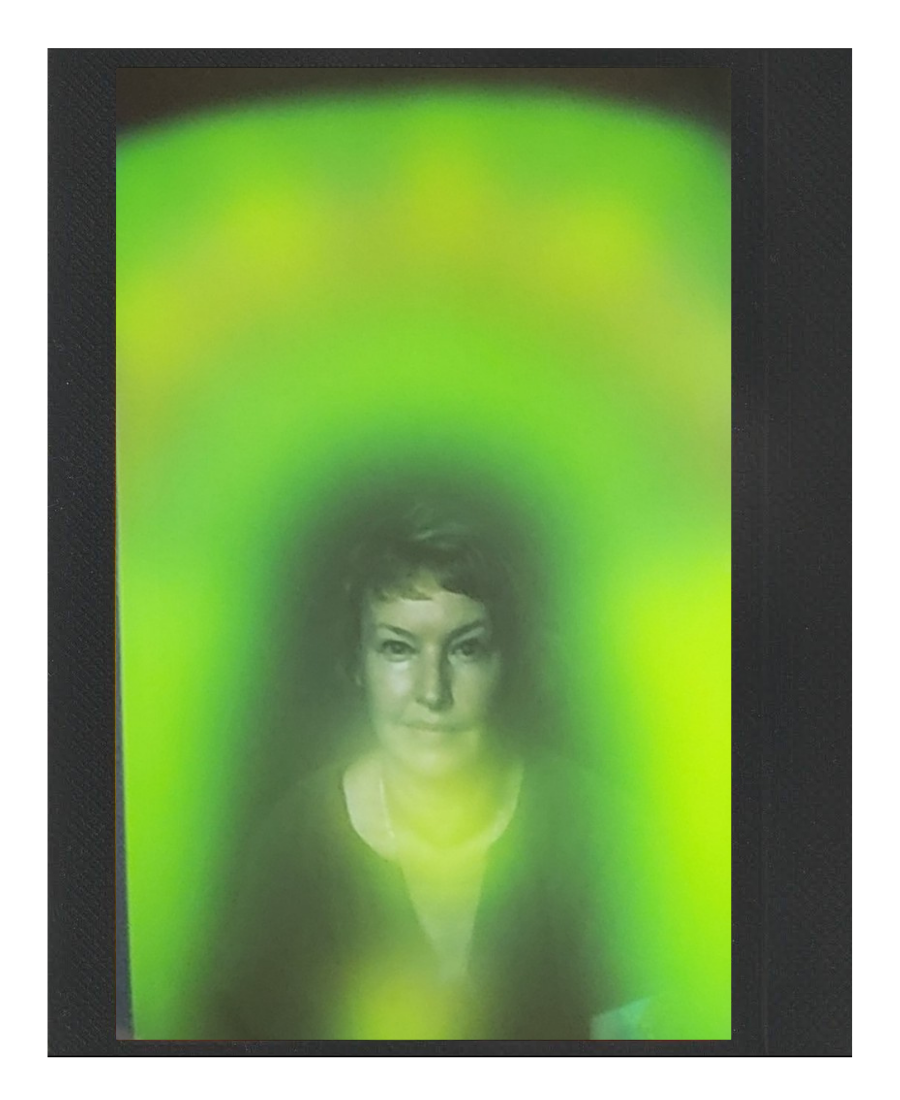 A woman’s face appears behind a green, misty screen or light projection with a black border.