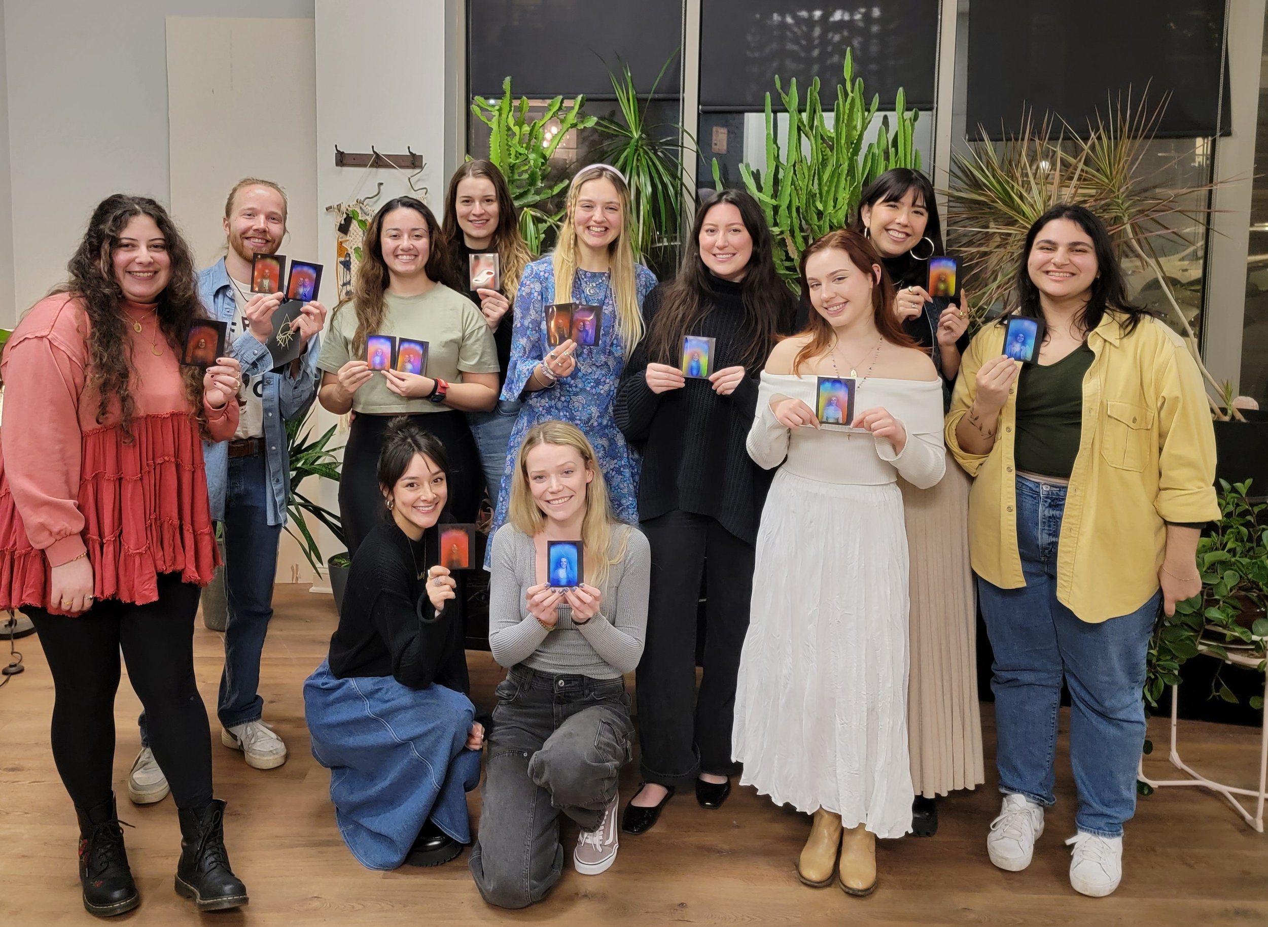 Group of ten diverse young adults smiling and holding tarot cards, standing in front of green plants in an indoor setting.