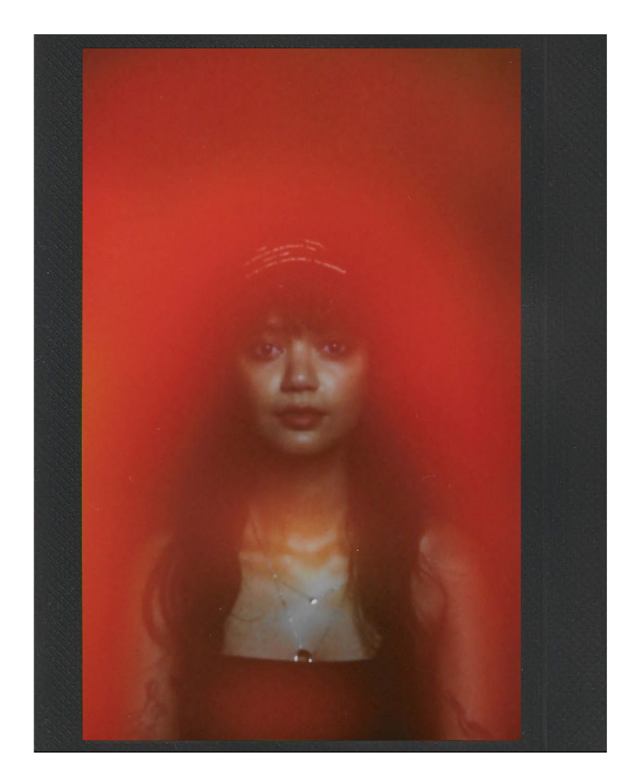 A woman with long dark hair, wearing a headband, a black tank top, and jewelry, looks directly into the camera against a red background.