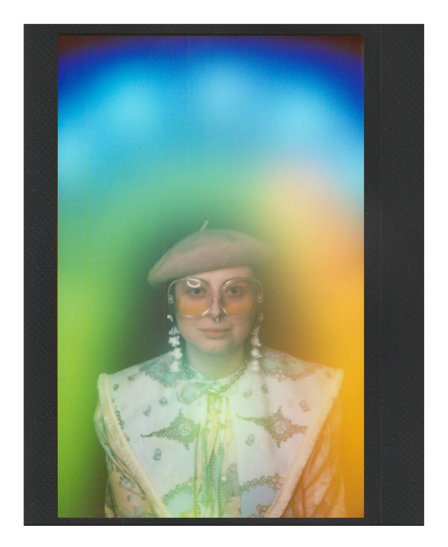 A person wearing large orange sunglasses, a beige beret, and vintage-style earrings, with a Western-style embroidered vest and a necktie, is seen through a rainbow-colored, blurred plastic sheet or window.
