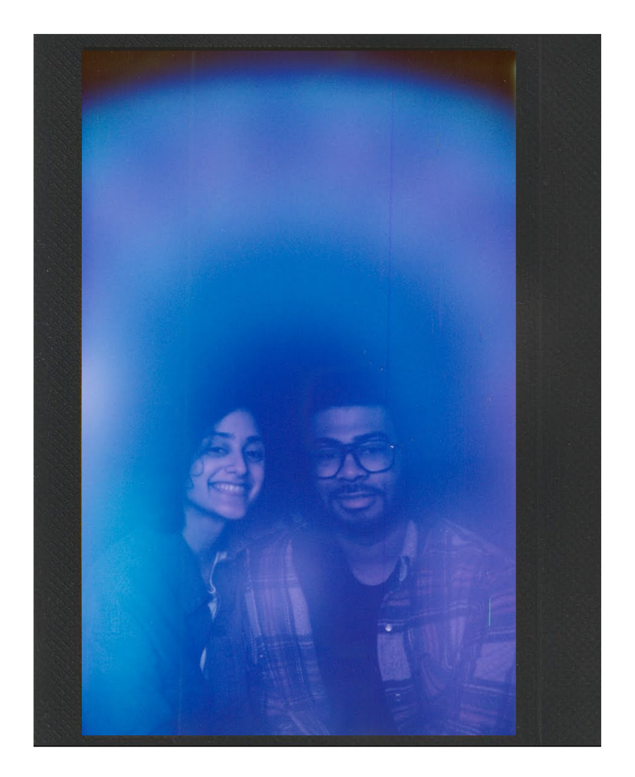 Two people taking a mirror selfie, obscured by a blue light or filter, with smiling woman on the left and man with glasses on the right.