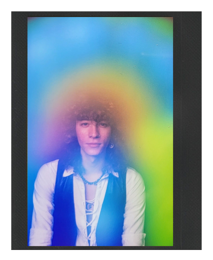 A person with curly hair wearing a white shirt, a black vest, and jewelry, standing behind a rainbow-colored transparent sheet, creating rainbow light reflections around their face.