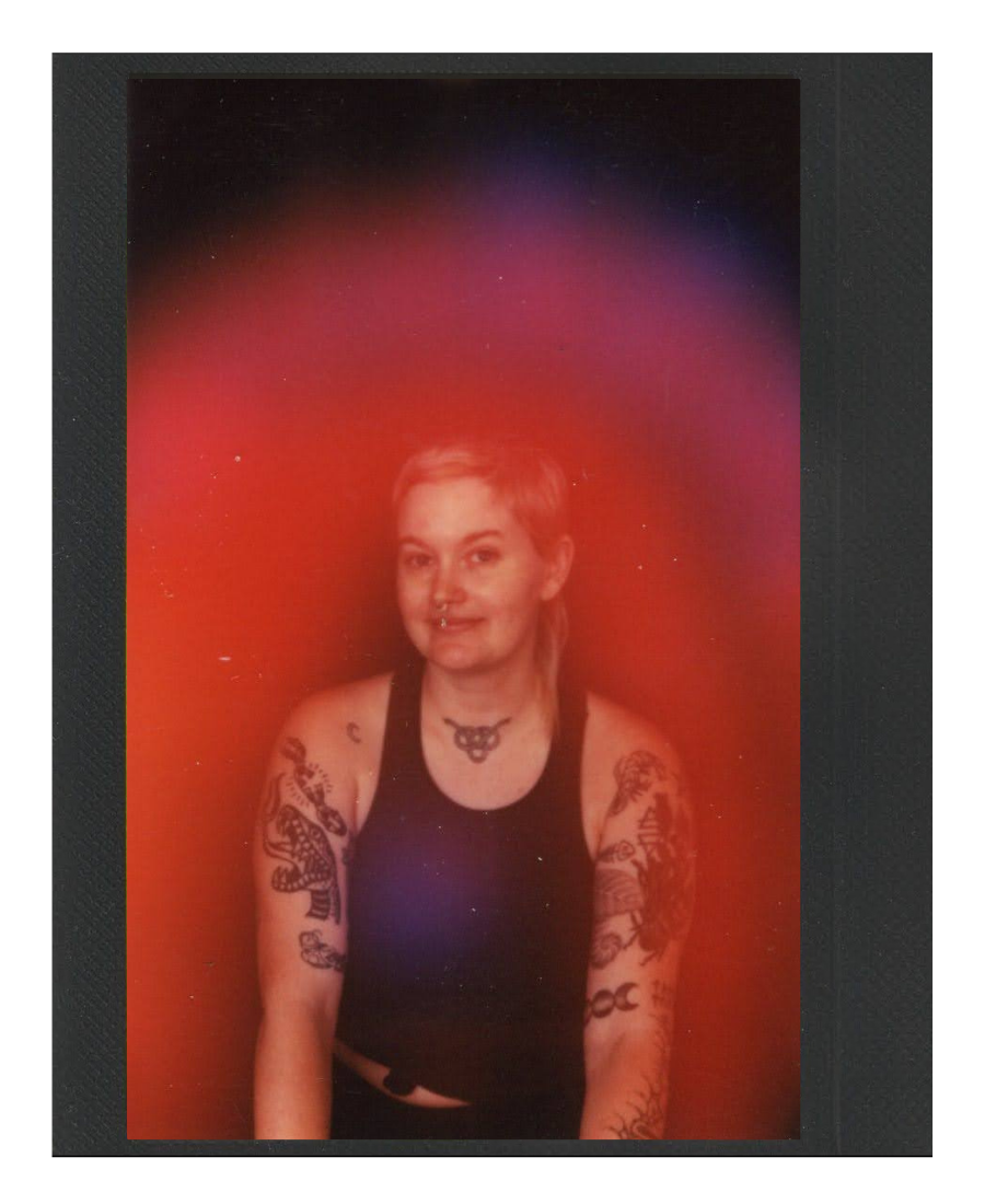 A woman with short, blonde hair and tattoos on her arms, wearing a black tank top and a choker necklace, standing against a colorful gradient background.