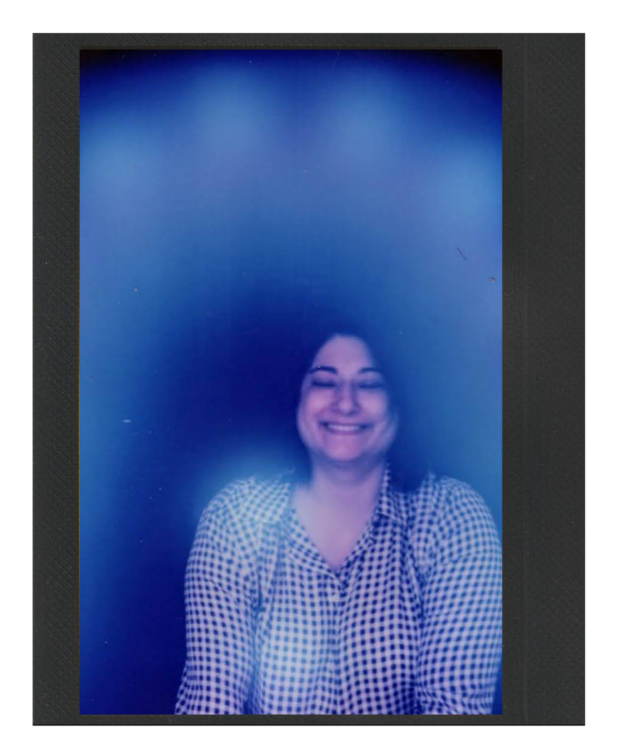A woman with shoulder-length dark hair and a big smile, wearing a checkered shirt, is sitting on a couch or chair against a dark background with a blue gradient glow around her.