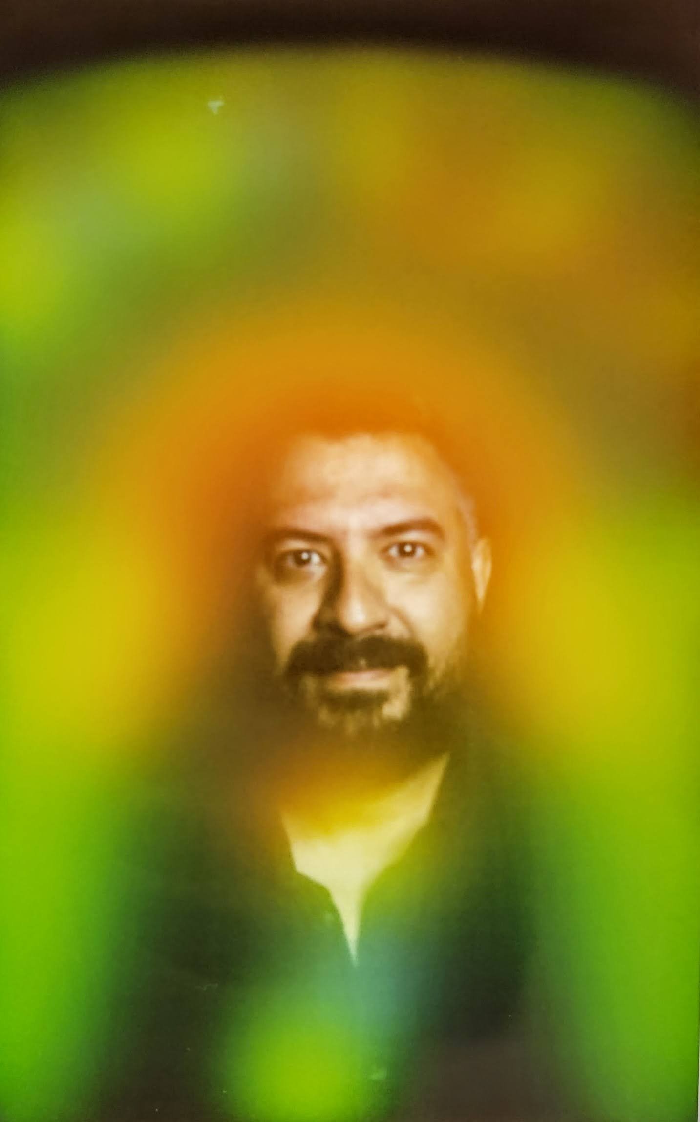A man with dark hair and a beard, wearing a black shirt, is seen smiling with a blurred green background.
