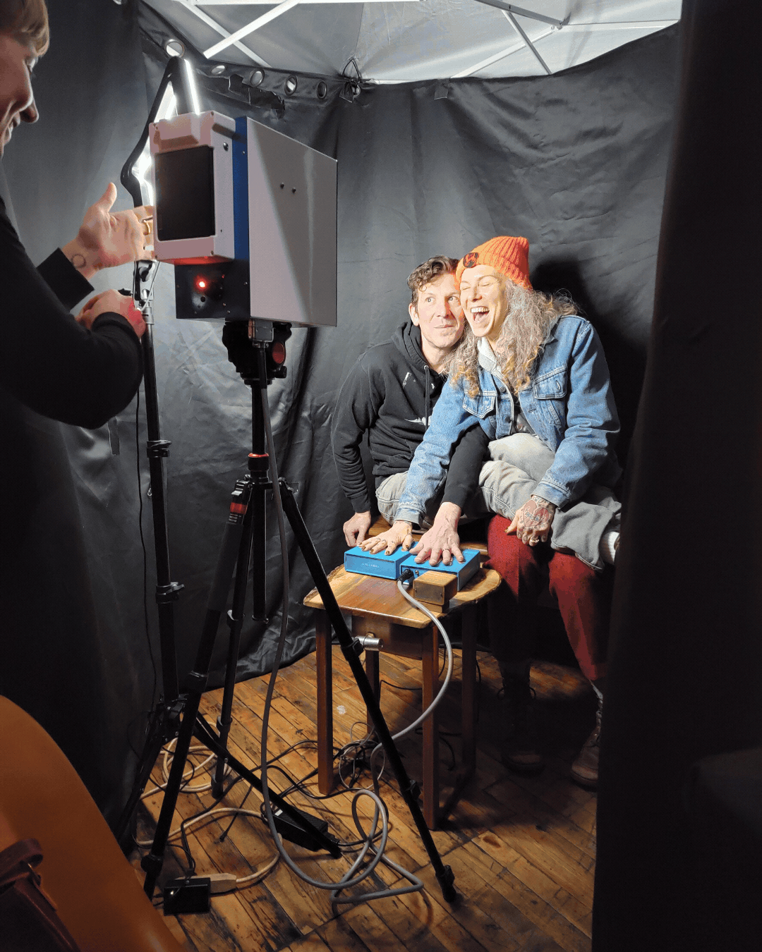 Two people posing for a photo in a dark photo studio, one wearing a denim jacket and orange beanie, the other in a black hoodie, with a photographer taking their picture using a vintage camera on a tripod.