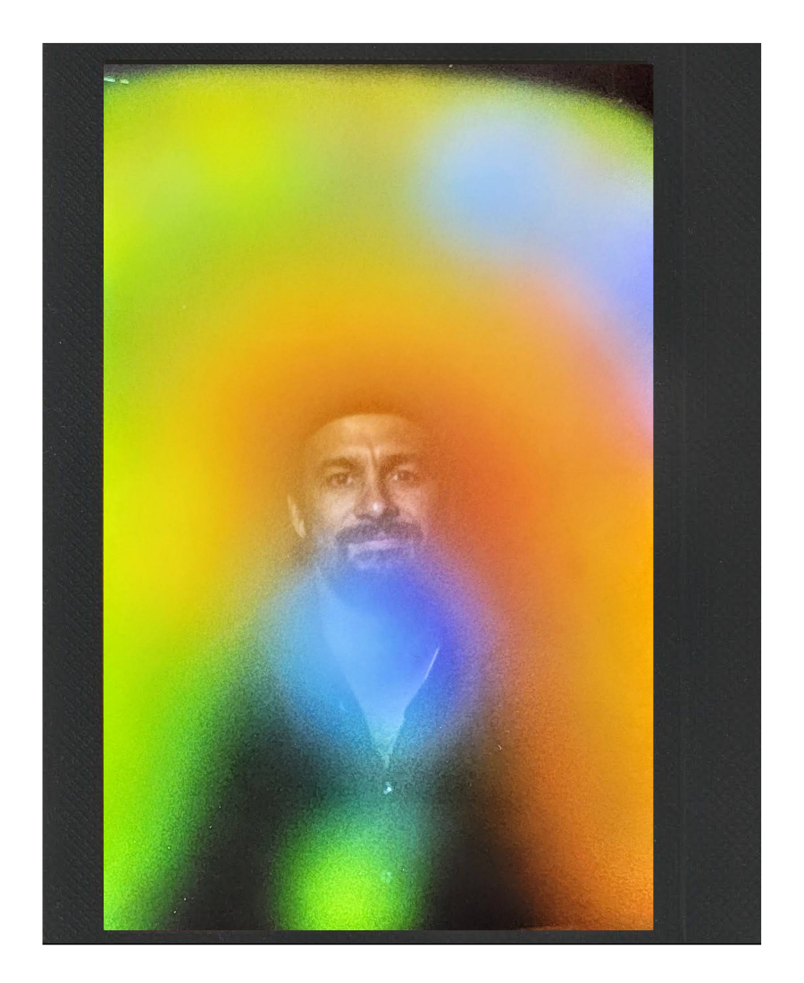 This image shows a man with a beard and mustache looking through a colorful, abstract rainbow-colored translucent surface or screen that creates a blurred, rainbow effect around his face.