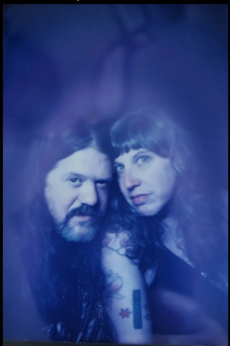 A blurry portrait of a man with long dark hair and beard, and a woman with curly dark hair, both looking at the camera, with a dark background.