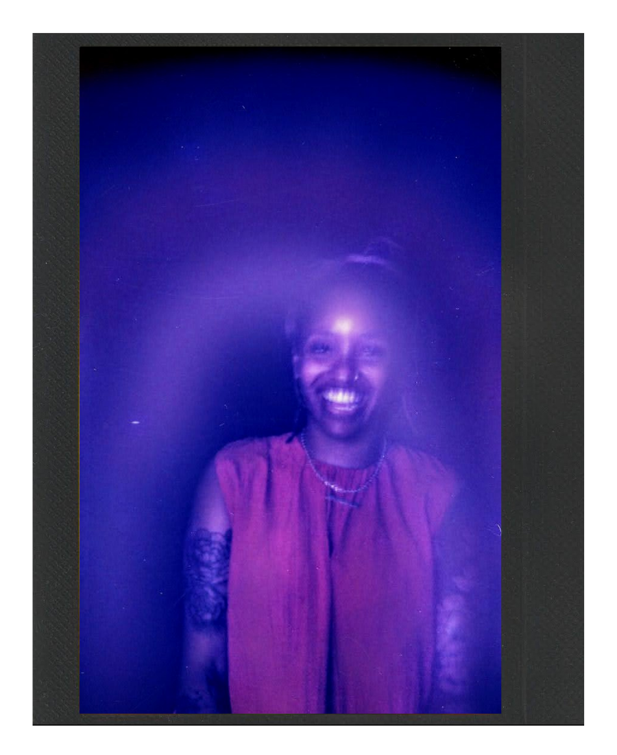 A person smiling in a purple-lit environment, wearing a sleeveless top with tattoos on the arms and a necklace.