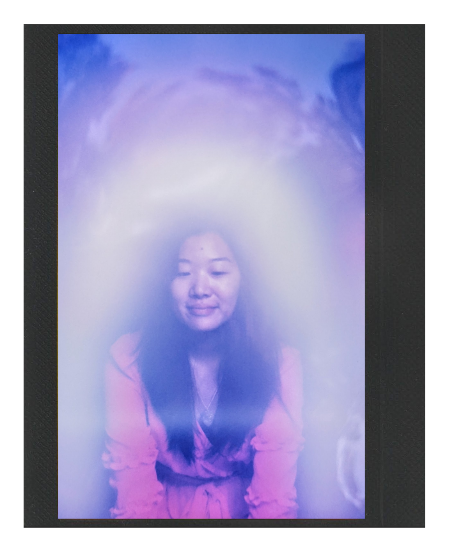 A young woman with long dark hair and closed eyes, wearing a pink top, is sitting with a foggy or misty purple and blue effect surrounding her, giving a dreamy atmosphere.