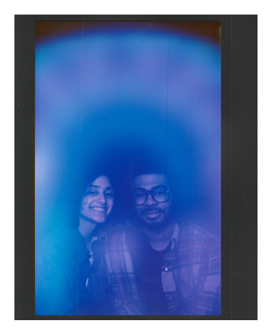 Two people take a selfie inside a photo booth with a blue-tinted background. The woman on the left has shoulder-length dark hair and is smiling, while the man on the right wears glasses and a plaid shirt.