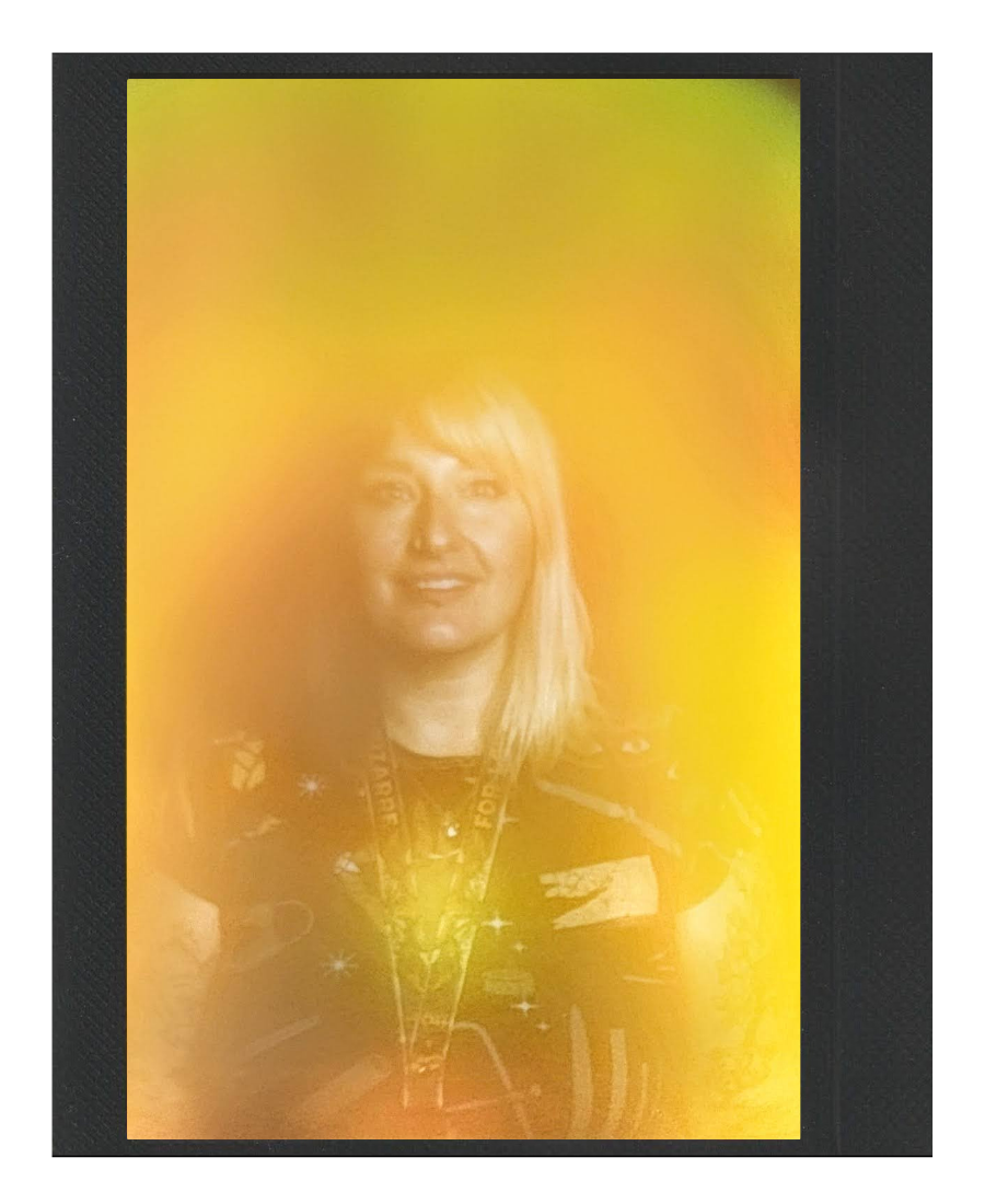 A woman with blonde hair smiling, wearing a galaxy-themed shirt and a lanyard around her neck.