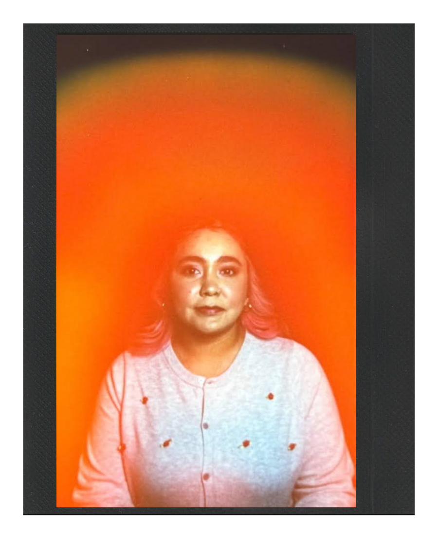 Portrait of a woman with light hair and makeup, wearing a light gray sweater with small black and red dots, against a vibrant orange and yellow background with a dark border.