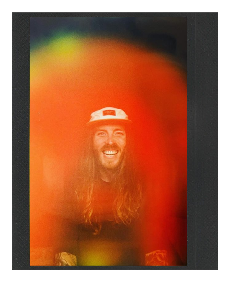 A man with long hair and a beard is smiling at the camera, wearing a white hat.