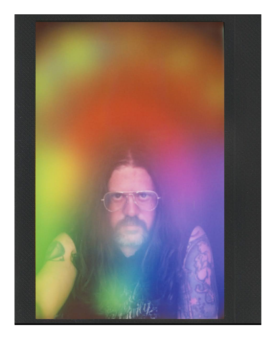 A person with long hair, glasses, and tattoos looks directly at the camera, surrounded by colorful rainbow haze.