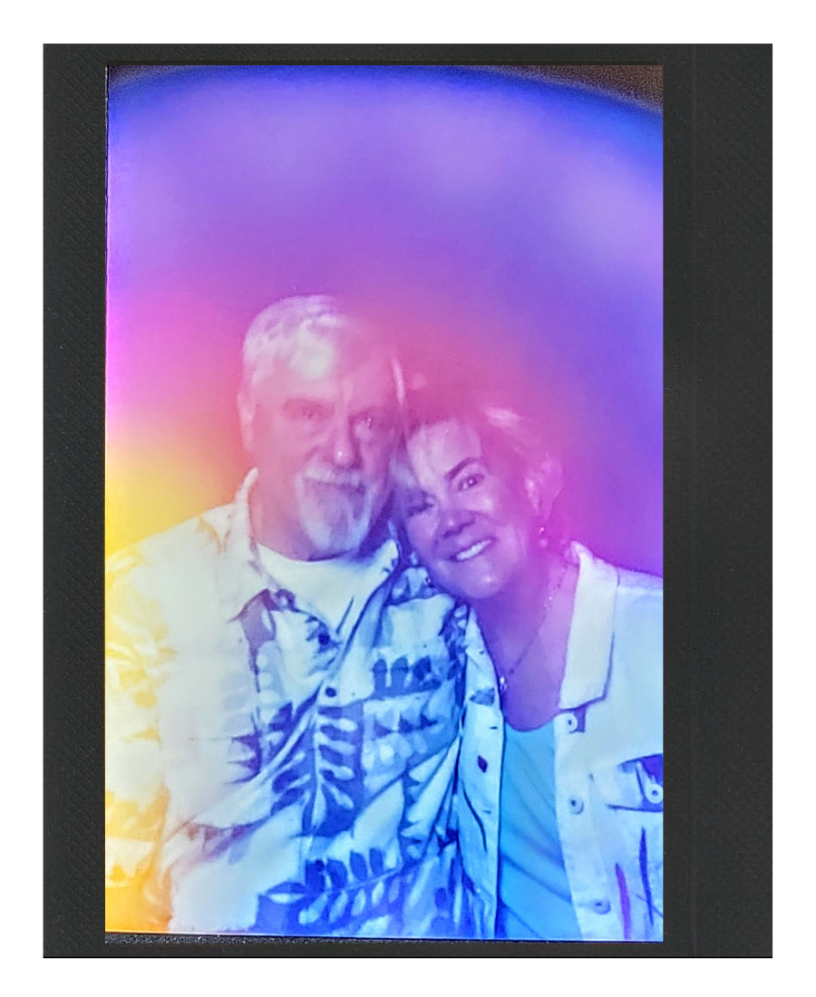 Colorful rainbow-like light overlay on a photograph of an older man with gray hair and beard and a woman with short hair, both smiling and posing together.