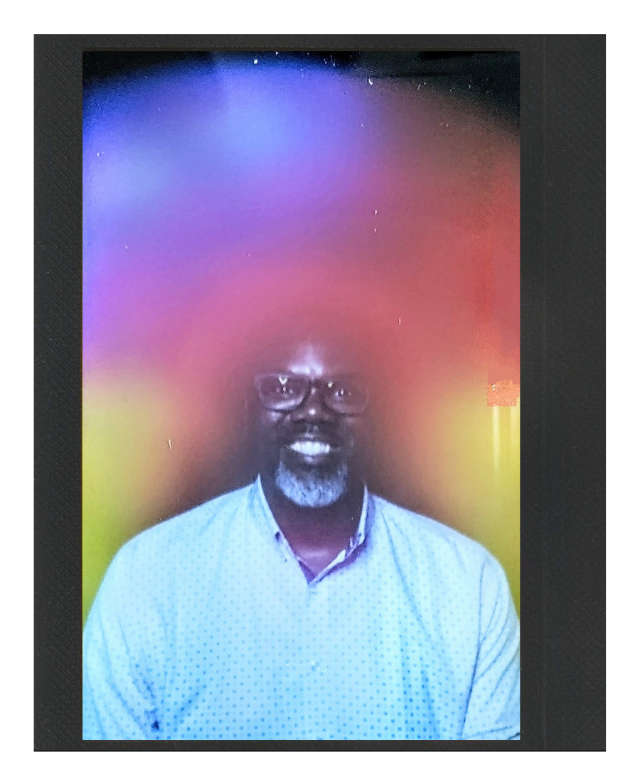 A man with glasses, a gray beard, and a mustache is smiling at the camera. He is wearing a light blue, button-up shirt with small polka dots, and the background features a colorful light display with fades of purple, yellow, and red hues.