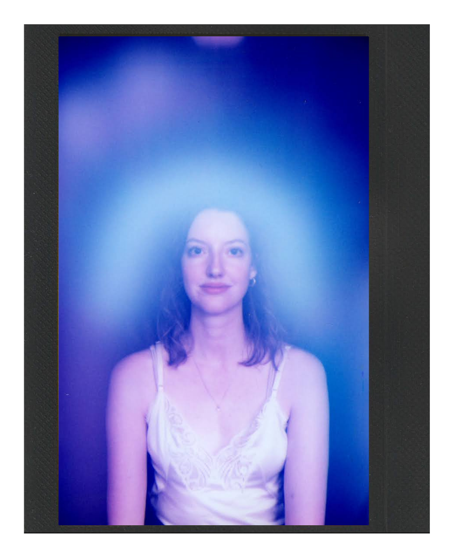 A woman with shoulder-length brown hair, wearing a white camisole top, stands in front of a vibrant, glowing blue background.