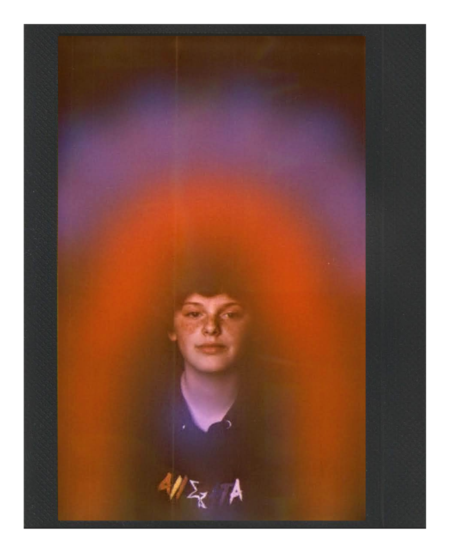 A person with short dark hair and freckled face looking at the camera, surrounded by a colorful, blurred, rainbow-like halo effect.