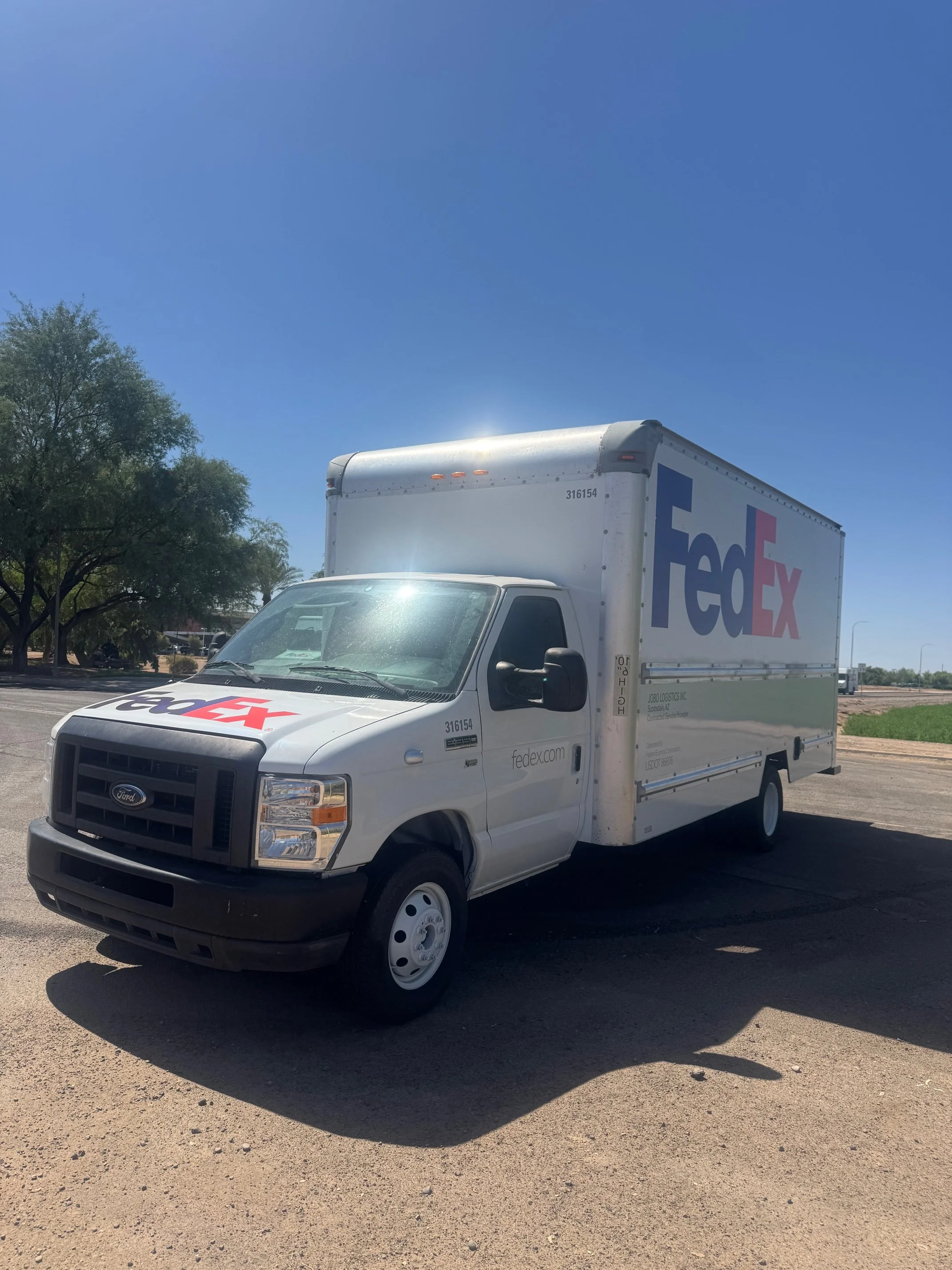 A white FedEx delivery truck parked on a paved lot under a clear blue sky, with some trees visible to the left.