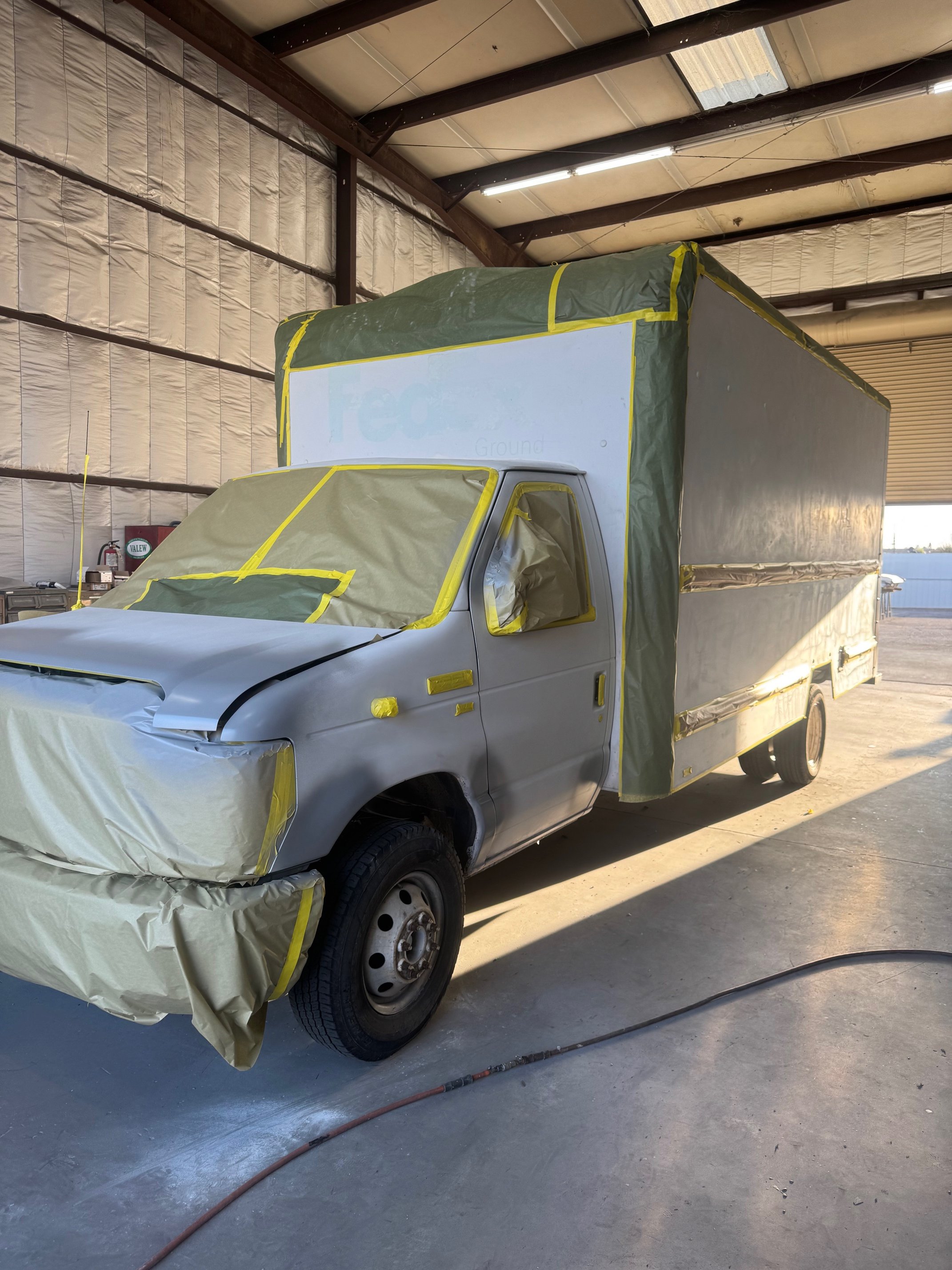 A silver pickup truck being coated with spray paint inside a large industrial warehouse. The truck is covered with masking paper and tape to protect areas from paint overspray.