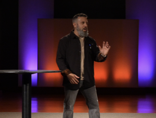 A man with a beard speaking on stage with purple and orange lighting in the background.