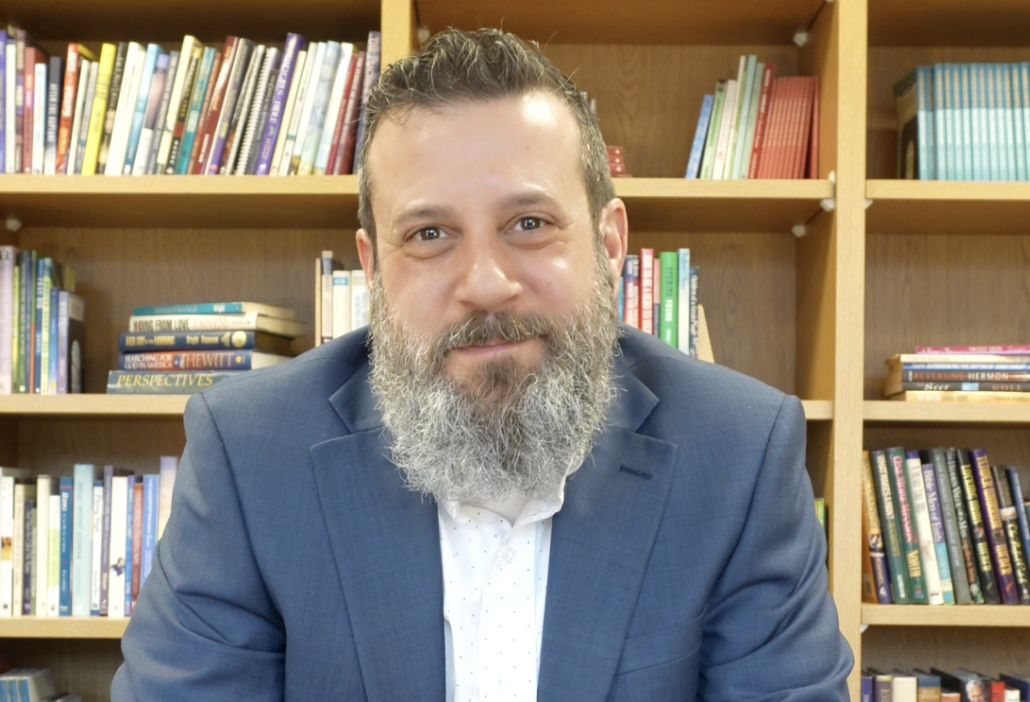 A man with a beard and short hair wearing a blue suit jacket, sitting in front of a bookshelf filled with various books.