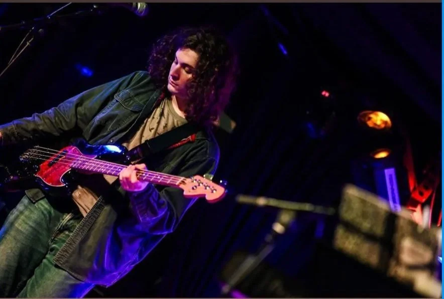 A person with curly hair playing an electric guitar on stage under purple and blue lighting.